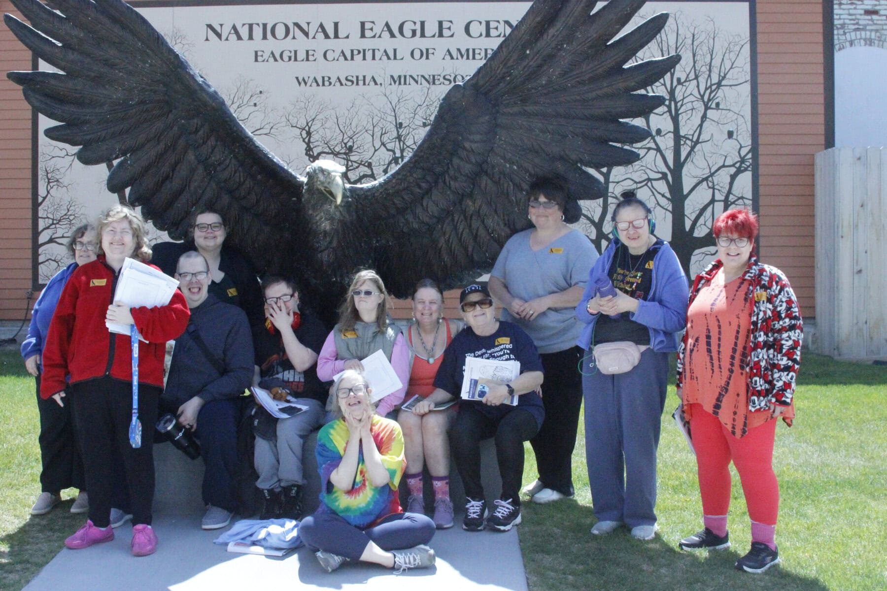 Eagle Center Group Photo 5-26 10 adults standing in front of a large eagle statue with its wings spread at the National Eagle Center