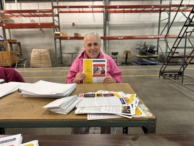 Getting the Winona ORC newsletter ready to mail a woman sitting at a table holding up the WORC newsletter with the table having parts of newsletters ready to be put together